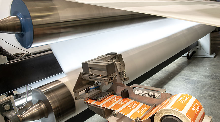 Automated label machine applying orange defect stickers to a roll of fabric